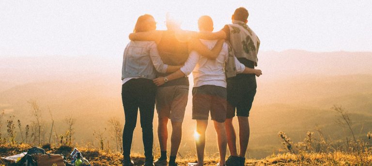 Vier Personen stehen umarmt vor einer sonnigen Landschaft bei Sonnenuntergang.